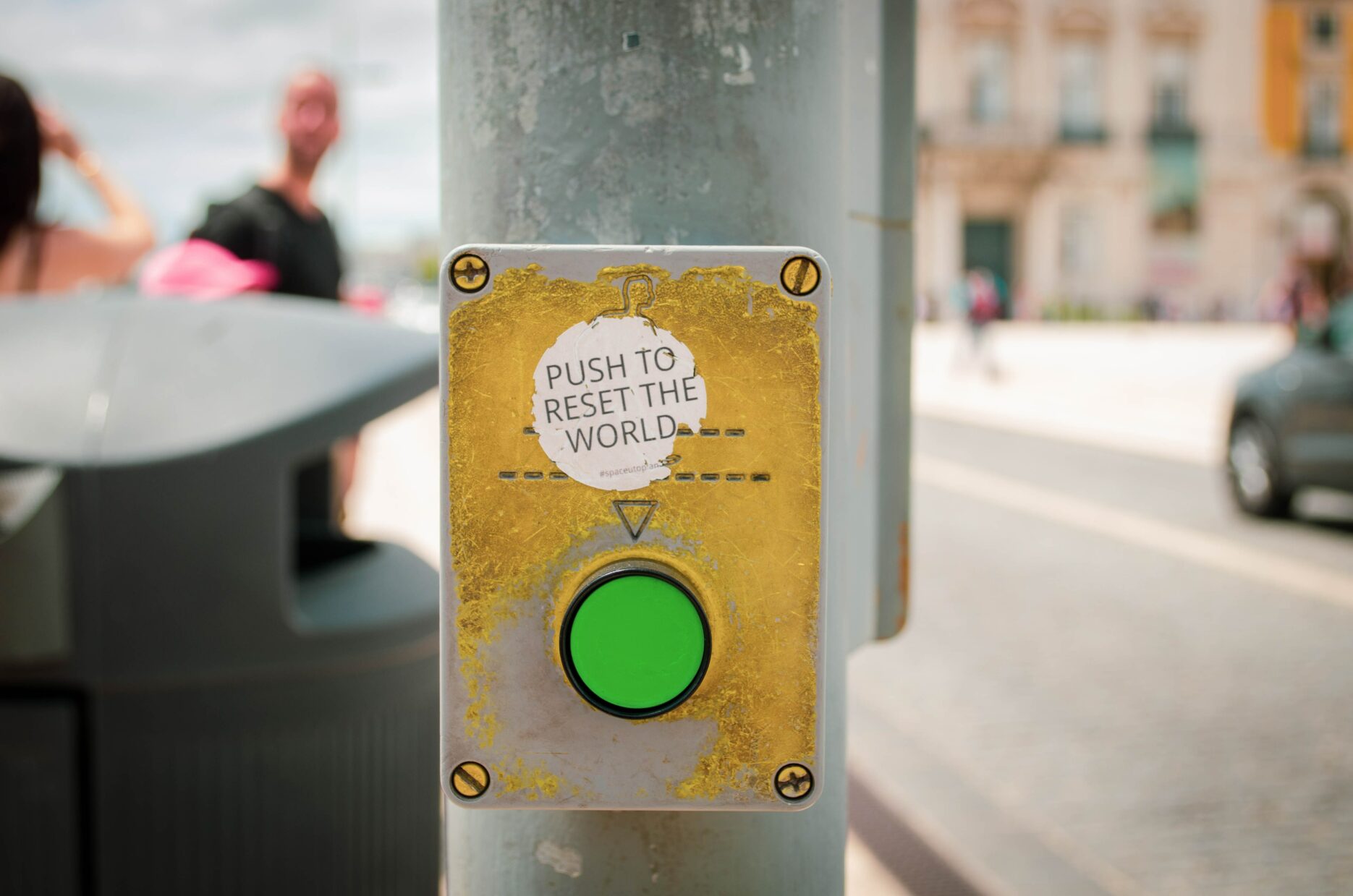 A pedestrian crossing button has been modified with a sticker that says, "Push to reset the world."