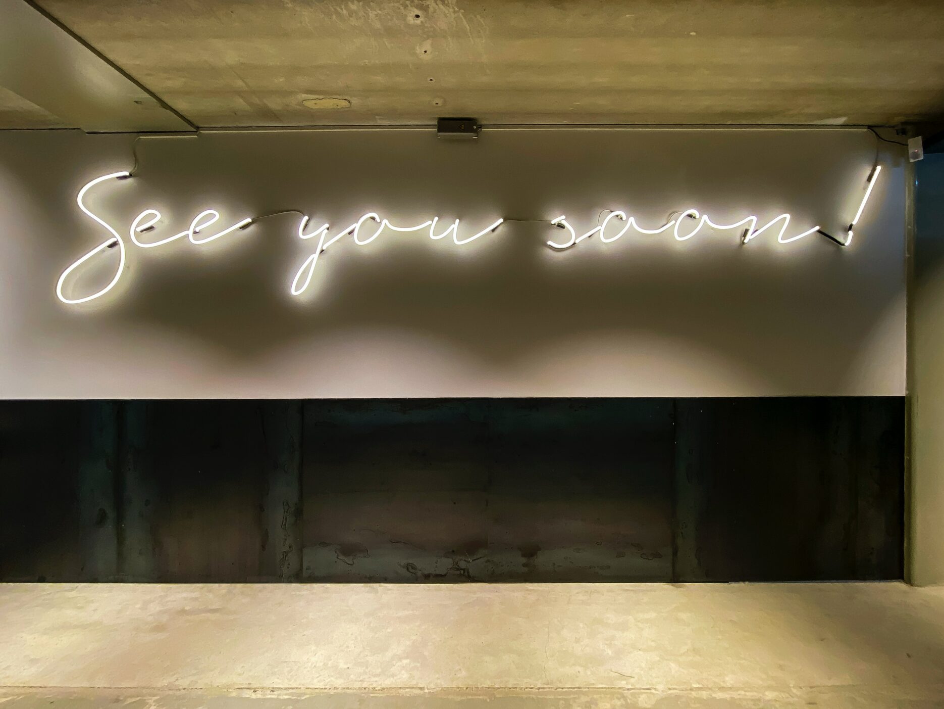 White neon script on a grey wall reads, "See you soon!"