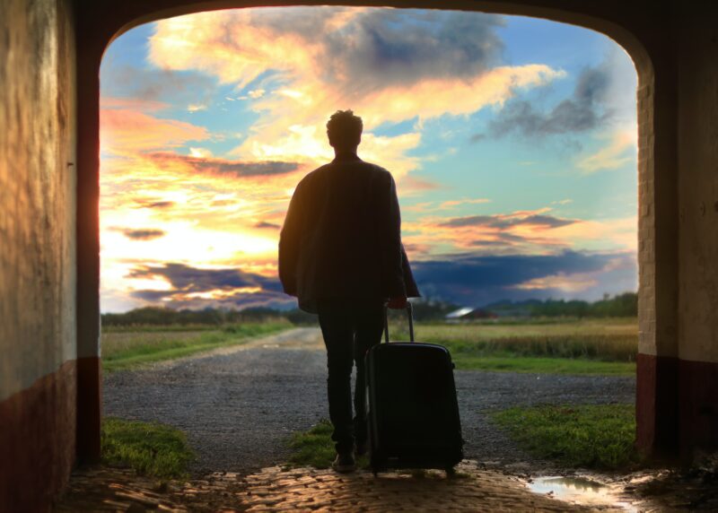 A young man framed in a doorway looking out into a sunrise. He is holding a large suitcase.