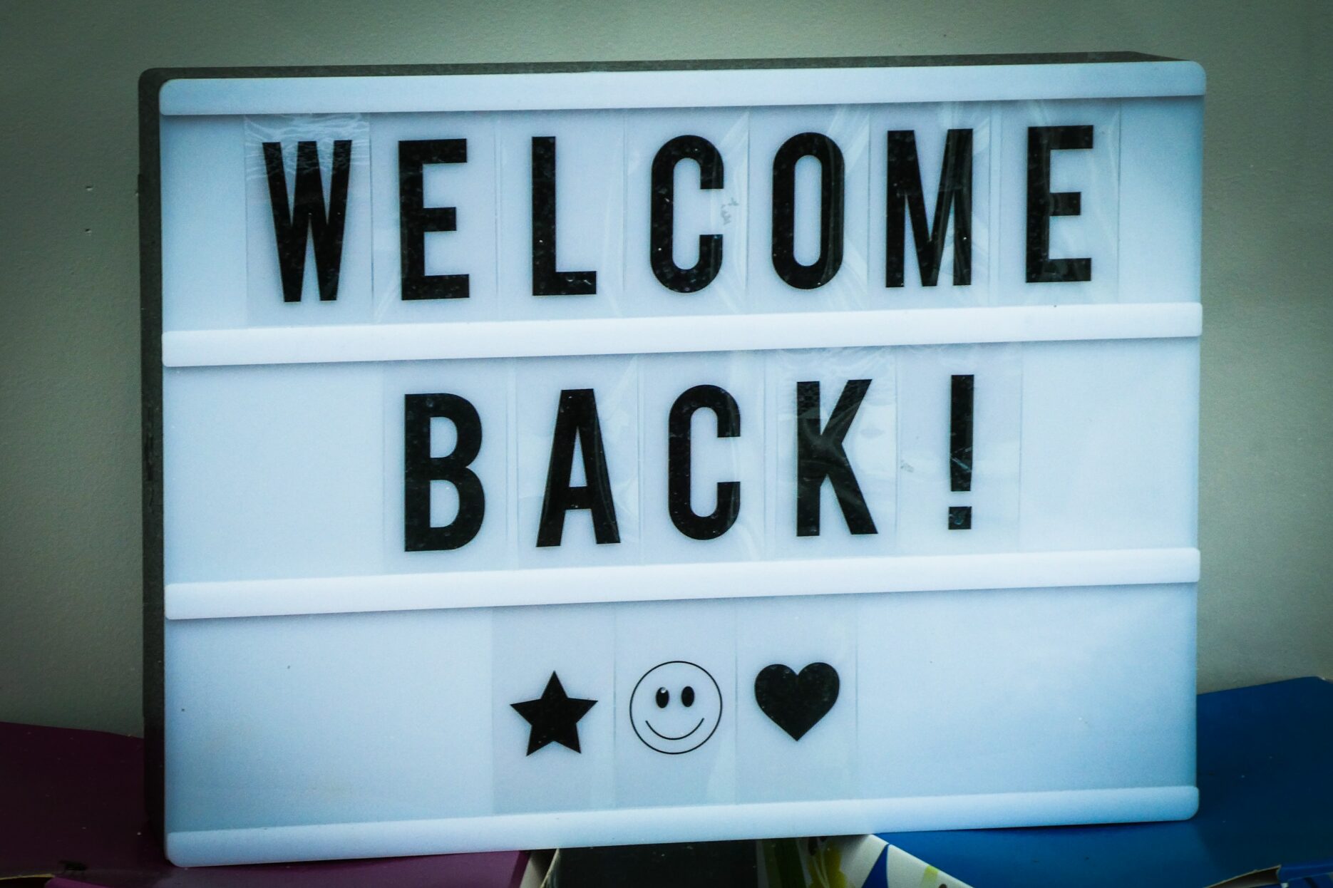 A white sign with removable black text reads "Welcome Back!" Below the words are a star, smily face, and heart. The sign is leaning against a wall and positioned on a desk.