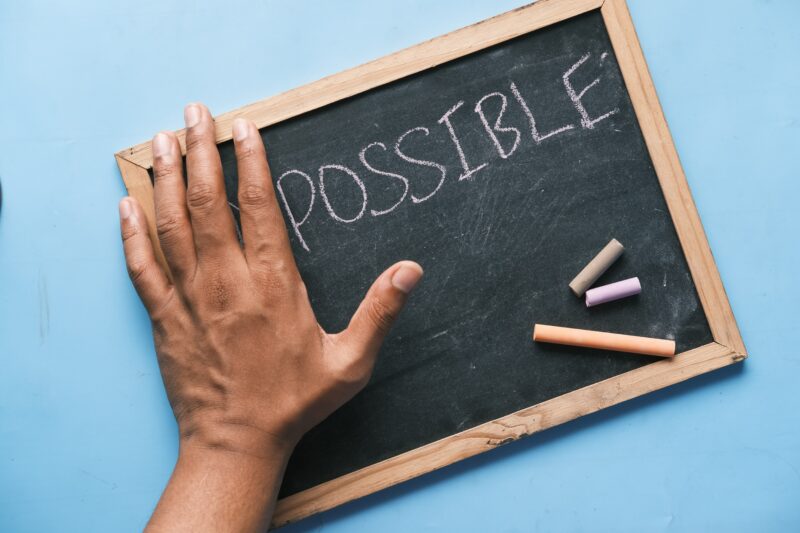 A chalkboard shows the word IMPOSSIBLE, but a hand covers the IM so it reads only POSSIBLE.