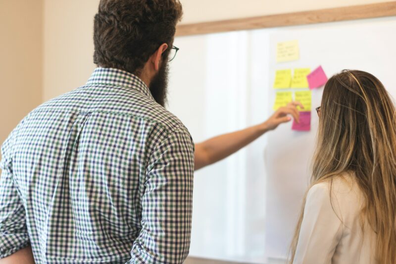 Two people look on as a facilitator places post-it notes on a white board.