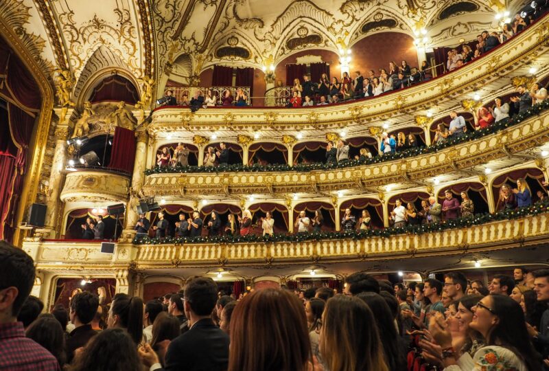 A crowd stands to applaud in an opulent theatre at the end of the performance.